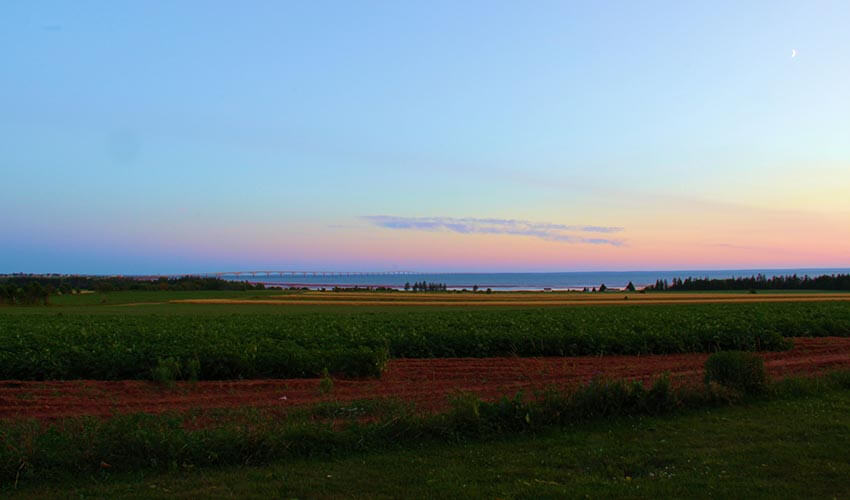 Farm fields in Prince Edward Island with Confederation Bridge and sunset in the background.