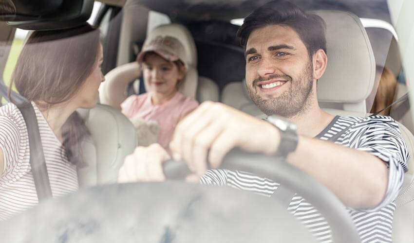 A happy family going on a road trip. Mother on passenger seat looking towards backseat at young child. Father is driving and smiling.