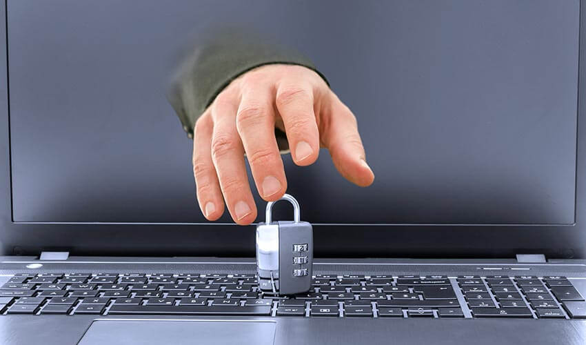 Closeup of hacker hand coming out of laptop screen trying to steal or break a padlock on keyboard.