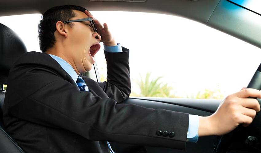 A man in a black suit yawning while driving a vehicle.