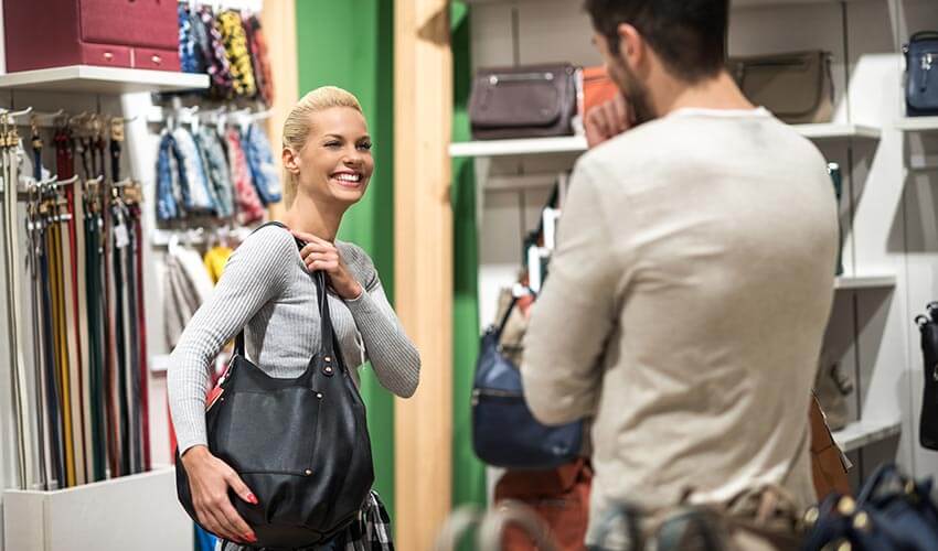 A man looking at a woman trying on a handbag in a store.