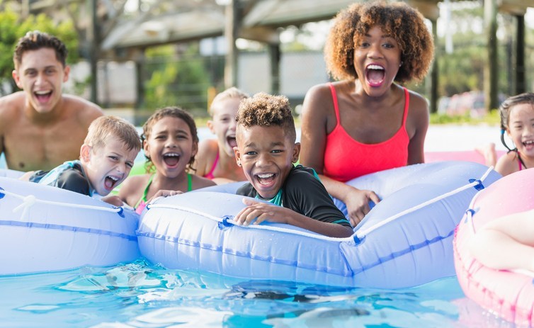 A group of multi-ethnic children and young adults on the lazy river at a water park, shouting with joy and looking at the camera. The focus is on the mixed race African American and Caucasian 9 year old boy in the center foreground.