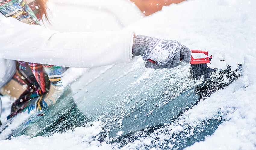 Woman clearing snow from windshield with a brush.