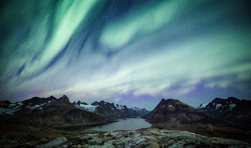 A man standing on rocks near a body of water looking at Northern Lights in the sky at night in East Greenland.