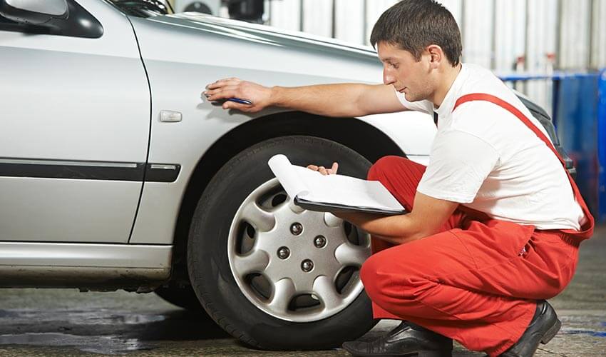 A mechanic squatting next to a silver car reading from a clipboard.