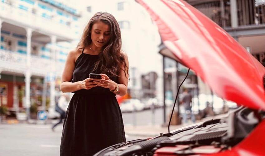 A woman standing next to a broken down car, making a call on her phone.