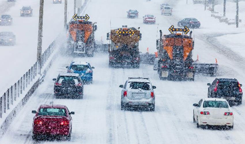 Three snow plow trucks clearing snow from a busy street with cars behind them.