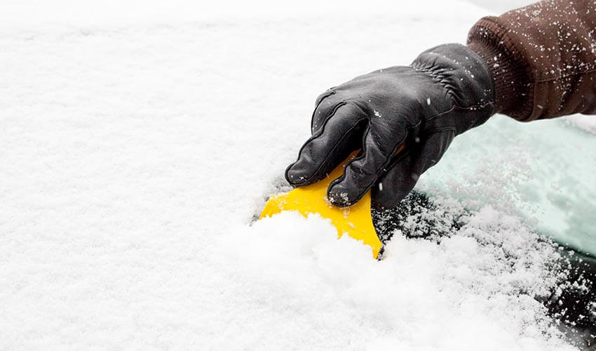A hand scraping ice off a windshield with a yellow ice scraper.