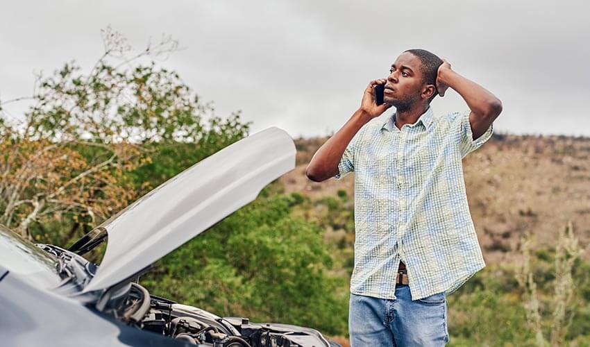 A young man calling roadside assistance while standing next to his broken down car.