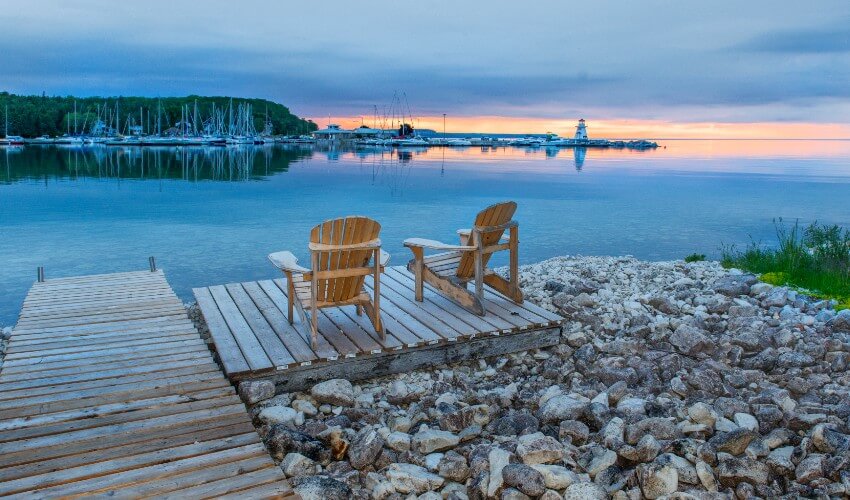 Two Adirondack chairs on a dock looking towards a marina and lighthouse at sunset.