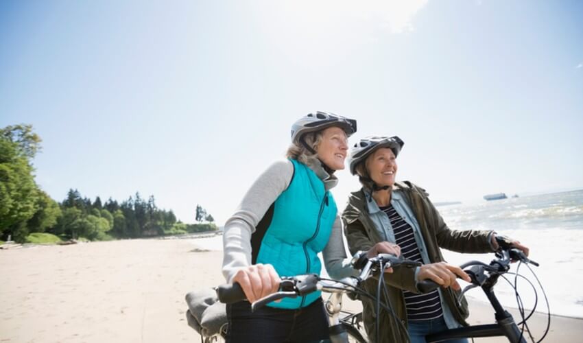 Two senior women walking with their bikes along a beach.