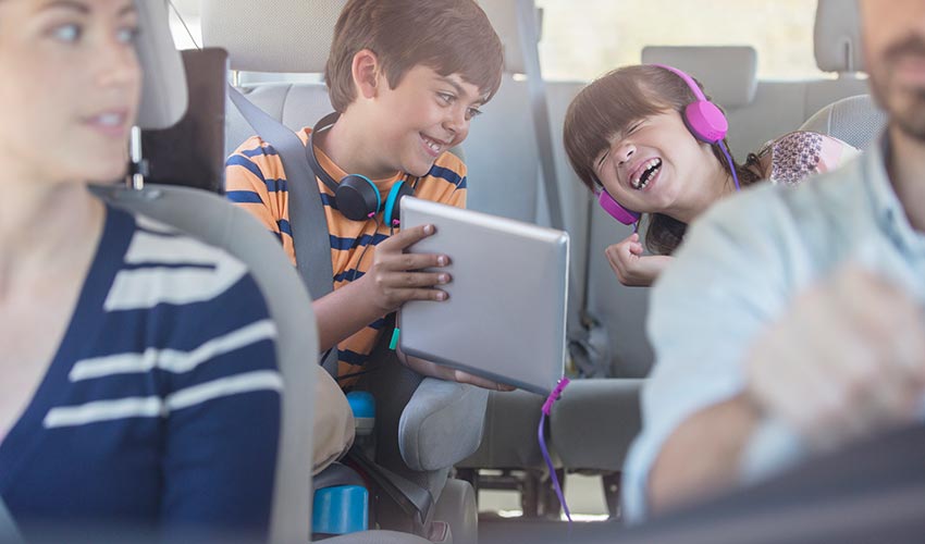 A family of four in a car. Kids are having fun in the back seat looking at a tablet.