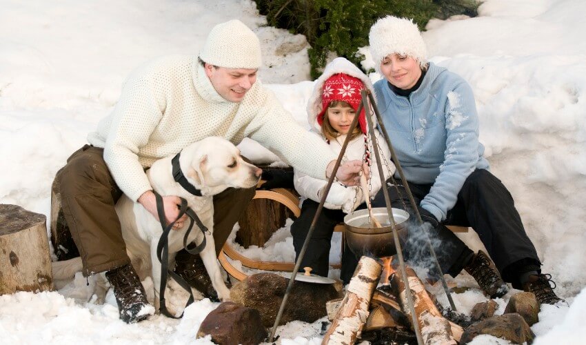 Family of three winter camping with their dog and cooking on a fire pit. 