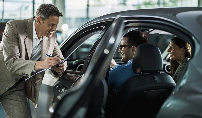 A car salesman taking notes and talking to young couple who is buying a car in a showroom.