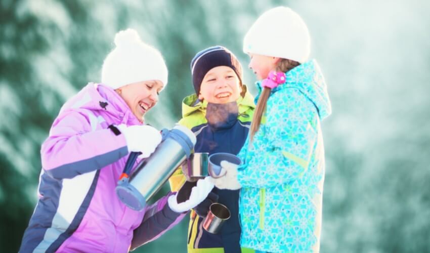 Mother and two children drinking hot tea near trees in a forest covered in snow.