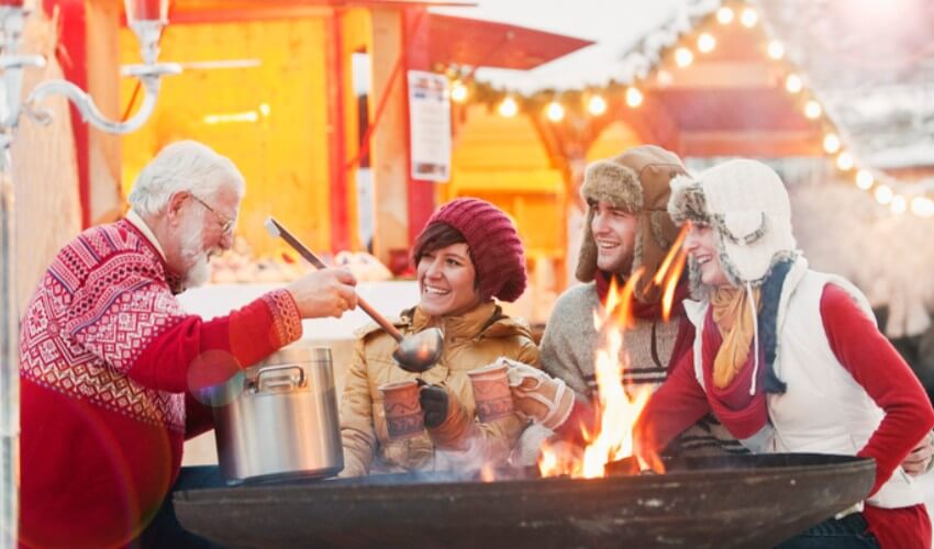 An older man pouring hot drinks for three visitors at a Christmas market.