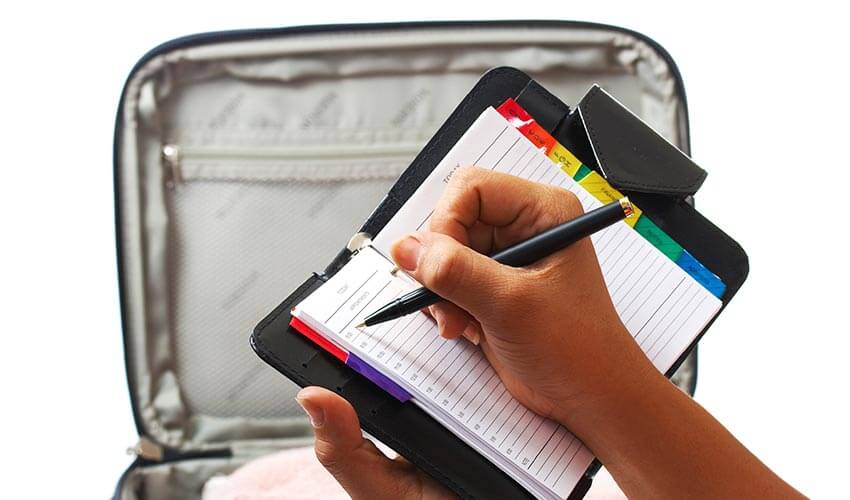 Writing out a packing list in a planner with suitcase in background.