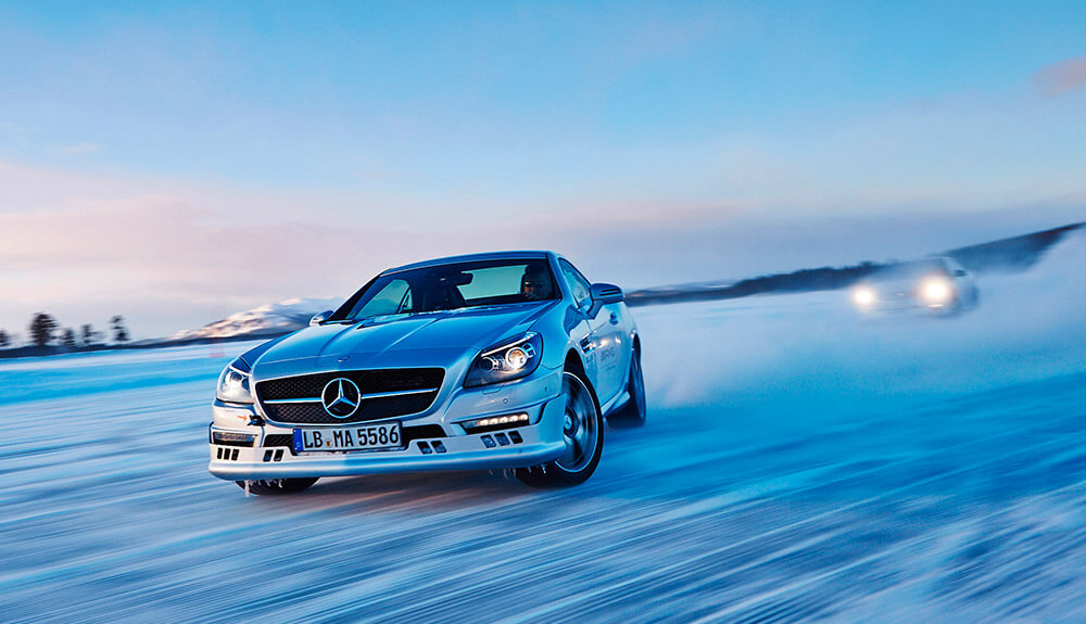 Silver Mercedes zooming down a snowy road, another car's headlights visible in the background behind