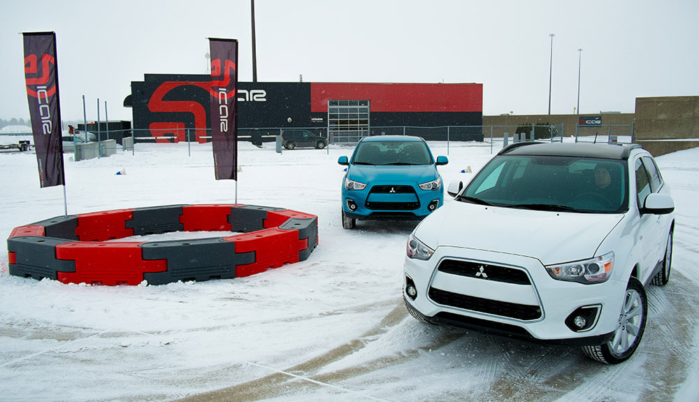 White and blue hatchbacks driving in a snowy roundabout, wheel marks visible in the ground and black and red flags with an ICAR logo