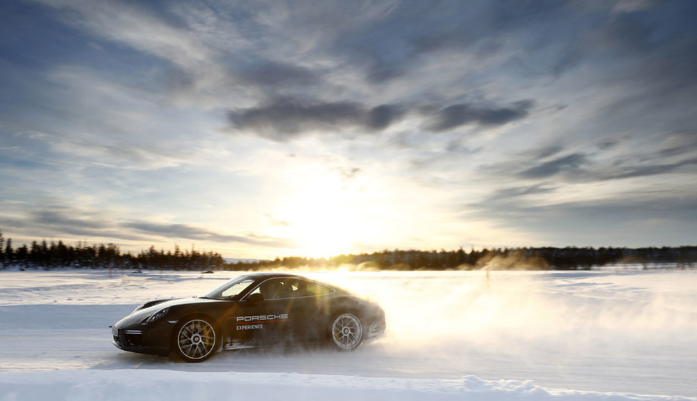 Black Porsche zipping down a snowy path with trees in the background, sun shimmering just behind the back wheels of the car
