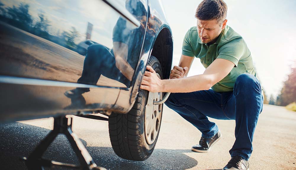 Man wearing jeans, sneakers and green t-shirt changing the tire on a black car
