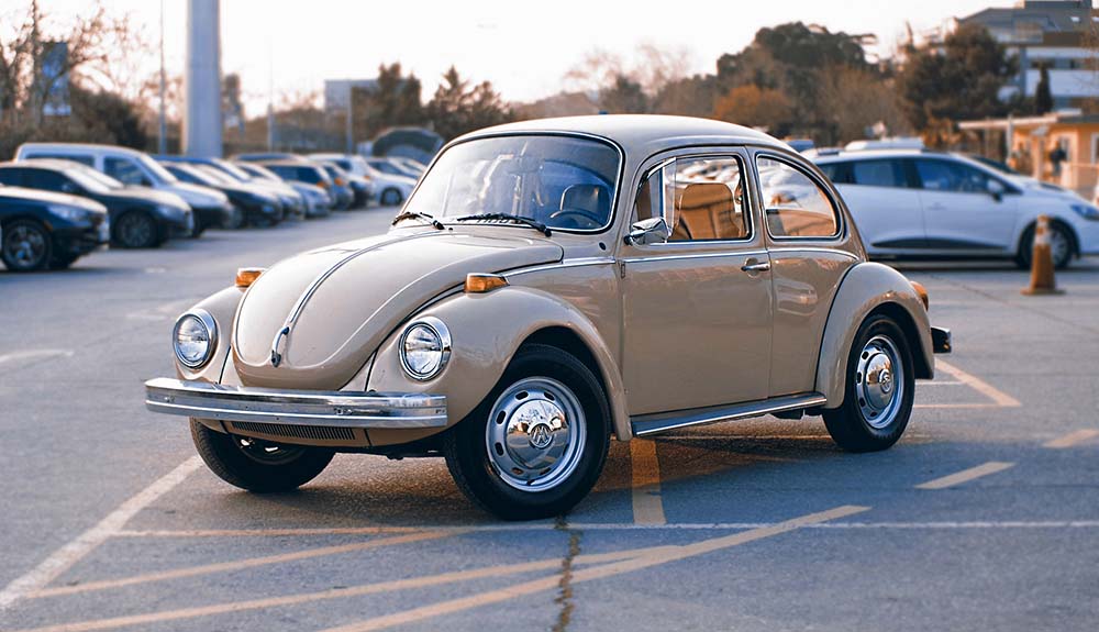 Light brown 1969 Volkswagen Beetle parked in busy parking lot at dusk, with various cars seen in the background