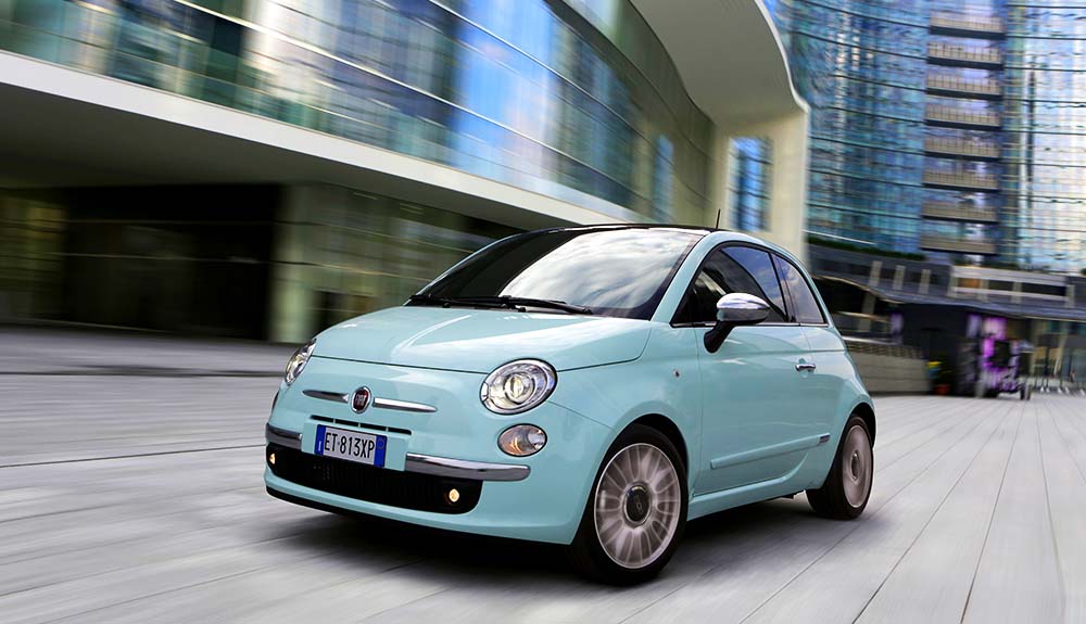 Light blue 2014 Fiat 500 Turbo zipping down urban road, with high rise buildings in the background