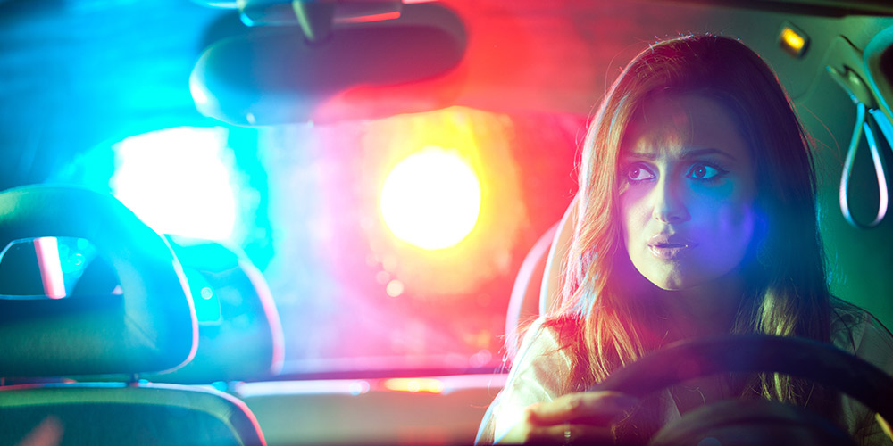 Police lights are visible through the rear window of a woman's car