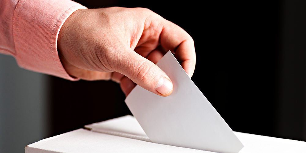 Person in salmon buttondown placing a closed ballot into an election ballot box