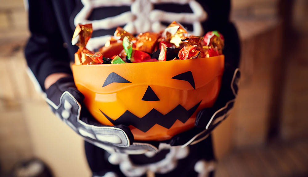 Child dressed as a skeleton holding an orange plastic bucket full of Halloween candy