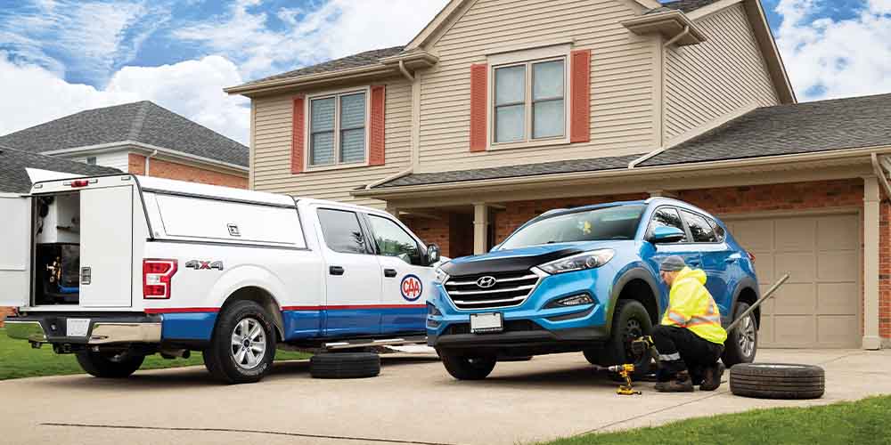 A CAA Roadside Assistance truck is shown in the driveway of a home, where a CAA mechanic changes the tire of a blue SUV