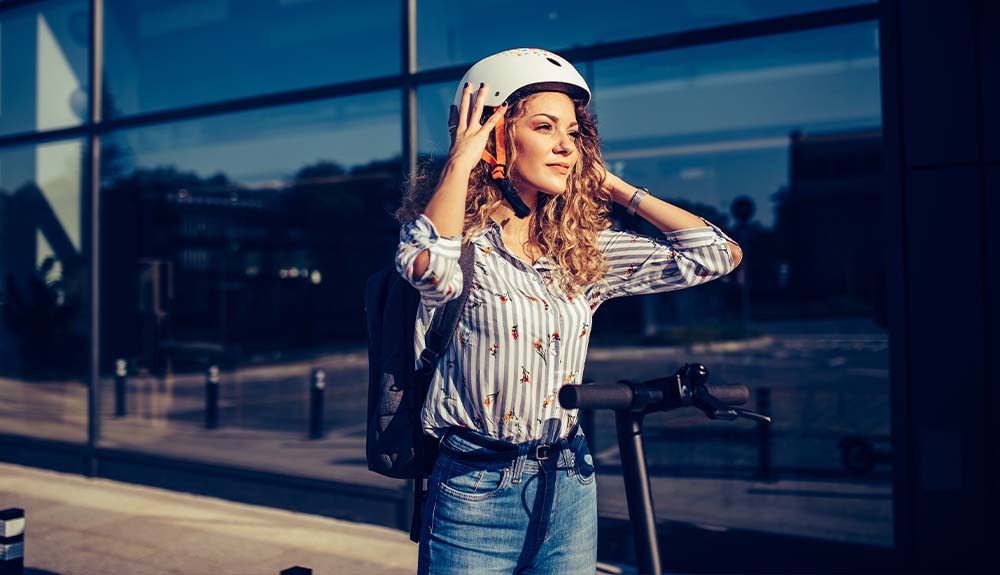A woman stands in front of a building adjusting her bike helmet