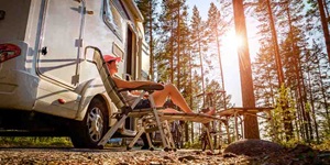 A woman sits on a lawn chair next to an RV in the woods