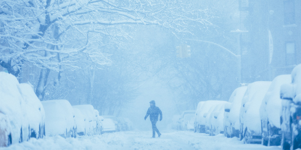 A person walking in the middle of a street covered in snow. Cars line the street that are also covered in snow, and there is snow falling.