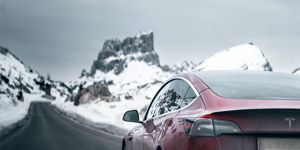 A red Tesla driving down a long road through snowy cliffs.