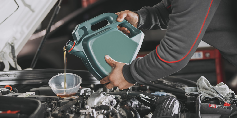 A person wearing a grey long sleeve sweater holds a green bottle of brown fluid over a car's open hood, pouring the liquid into a funnel leading into a part of the car's engine.