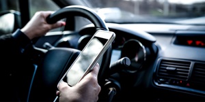 A close up view of a person with one hand on a steering wheel, and in their other hand they are holding a cellphone. 