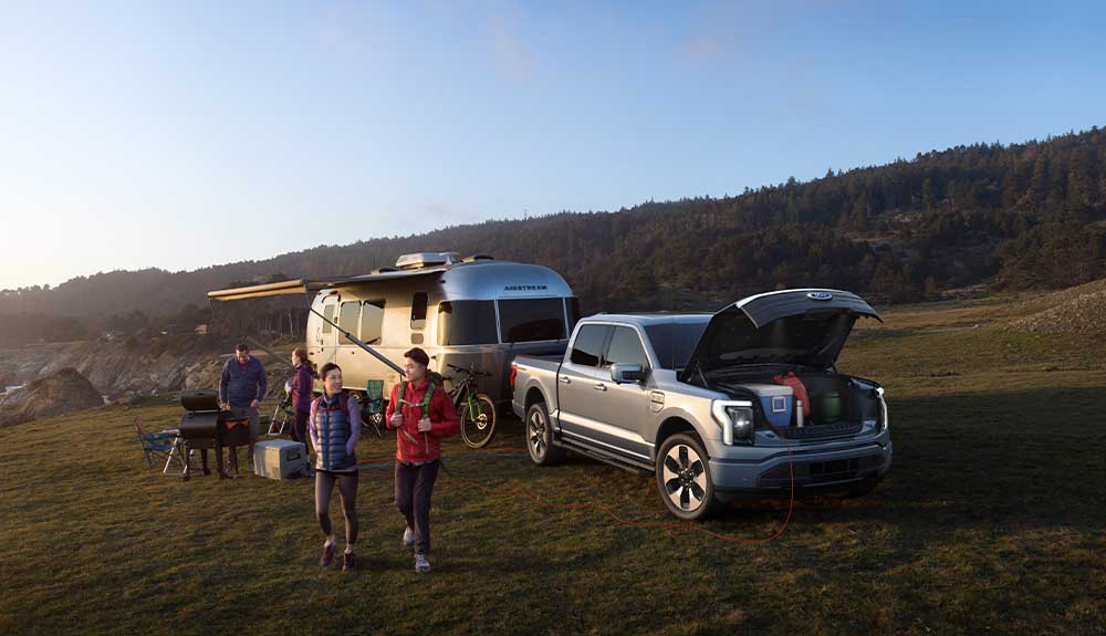 A silver truck with a camper hooked to the back of it is parked in a field with mountains behind it. There are two people in the background standing in front of a barbecue. There are two people in the foreground wearing vests and a backpack. The hood of the truck is open with a blue cooler and other camping gear is visible.
