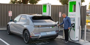 A man wearing a blue button-up shirt and black pants is standing at an EV charging station, which is white with green. He is holding a black hose up to his car to charge it.