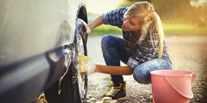 A woman squatting next to the rear wheels of a car holding a soapy yellow sponge scrubbing at the rims of a tire with a pink bucket next to her.