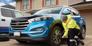 A man wearing a grey toque and a yellow safety sweater with the CAA logo is kneeling next to a blue SUV. He is holding a wrench with his hands that is positioned on the front driver tire of the car. Next to the SUV on the driveway is a white truck.