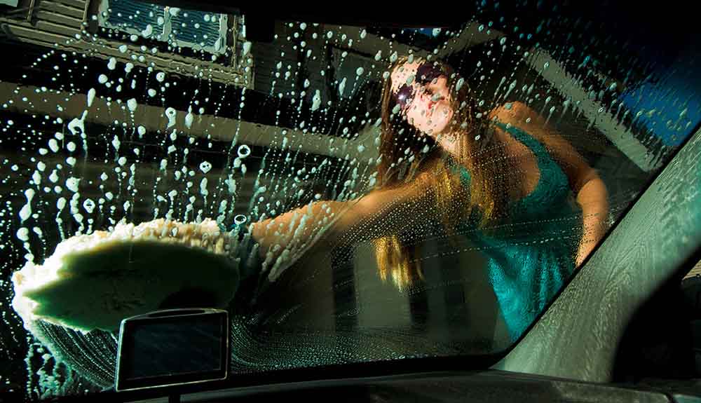 Woman with sunglasses and green tank top washing a soapy windshield with a sponge