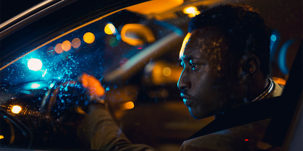 A man sitting in a car with one hand on the wheel looking through his front window. It is dark outside and there are spots of orange light reflected in his front dash.