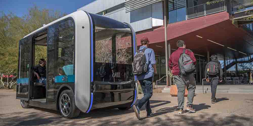 A box-shaped vehicle with wheels and its doors open and a single passenger sitting inside at the back. There are three people with backpacks on walking in front of the shuttle, which is stopped in front of them waiting for them to finish crossing.