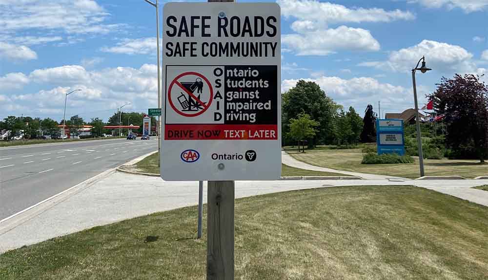 A sign on a wooden post that reads Safe Roads Safe Community. Below it is a black and white drawing of a martini glass, a cellphone, a marijuana leaf and keys that is crossed out with a circle in red. Down the side there are black letters that spell out the acronym OSAID, which stands for Ontario Students Against Drunk Driving. Below it in a red box is black copy that reads Drive Now Text Later. There is a CAA logo and an Ontario logo at the bottom of the sign.