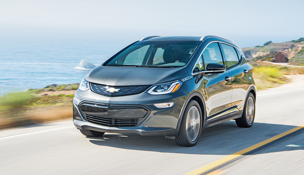 Gray Chevrolet Bolt EV driving down a winding road by a lake