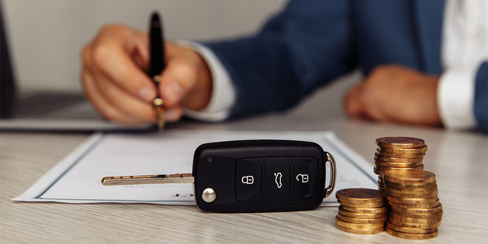 A black and silver car key with three buttons sits on top of a table next to a stack of loonies. Behind you see a contract on the table being signed by a person wearing a white shirt and blue blazer on top. They are holding a black and gold pen and you see only their arms and torso. 