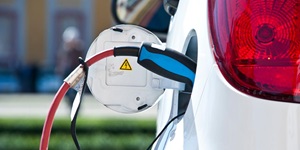 A close up of an electric vehicle charger plugging into the left side of a white vehicle. The charging port door on the vehicle is open and has a small yellow triangle sticker with the charging symbol on it. The charger is black with a blue stripe and a red cable. You can see the red tail light of the car. 