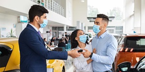 A man in a navy suit wearing a mask is handing over a small black item to another man wearing a button up blue shirt, who is also wearing a mask. There is a woman wearing a white striped button up shirt with her arms around the man in the blue shirt. Behind them is a car showroom.