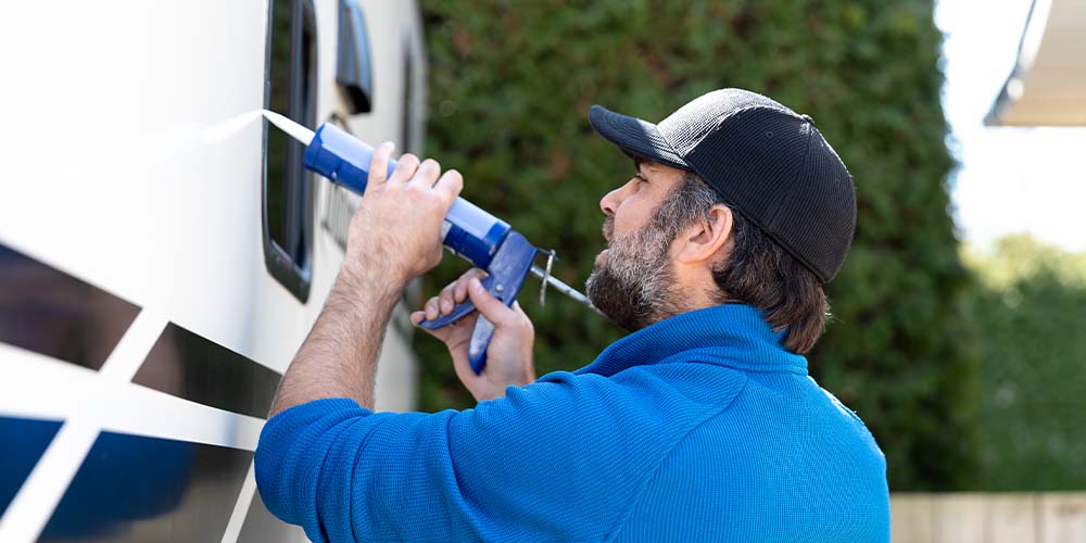 A side view of a man with a beard, wearing a black baseball cap and a bright blue shirt with his sleeves rolled up. He is holding a dark blue caulking gun with a pointed white tip up to a window on the side of a trailer, which is white with black stripes at the bottom.
